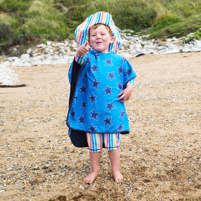 A young boy on a beach wearing a 'Beach Towel Poncho Blue Starfish', pointing his finger. He has a matching striped hat and shorts.